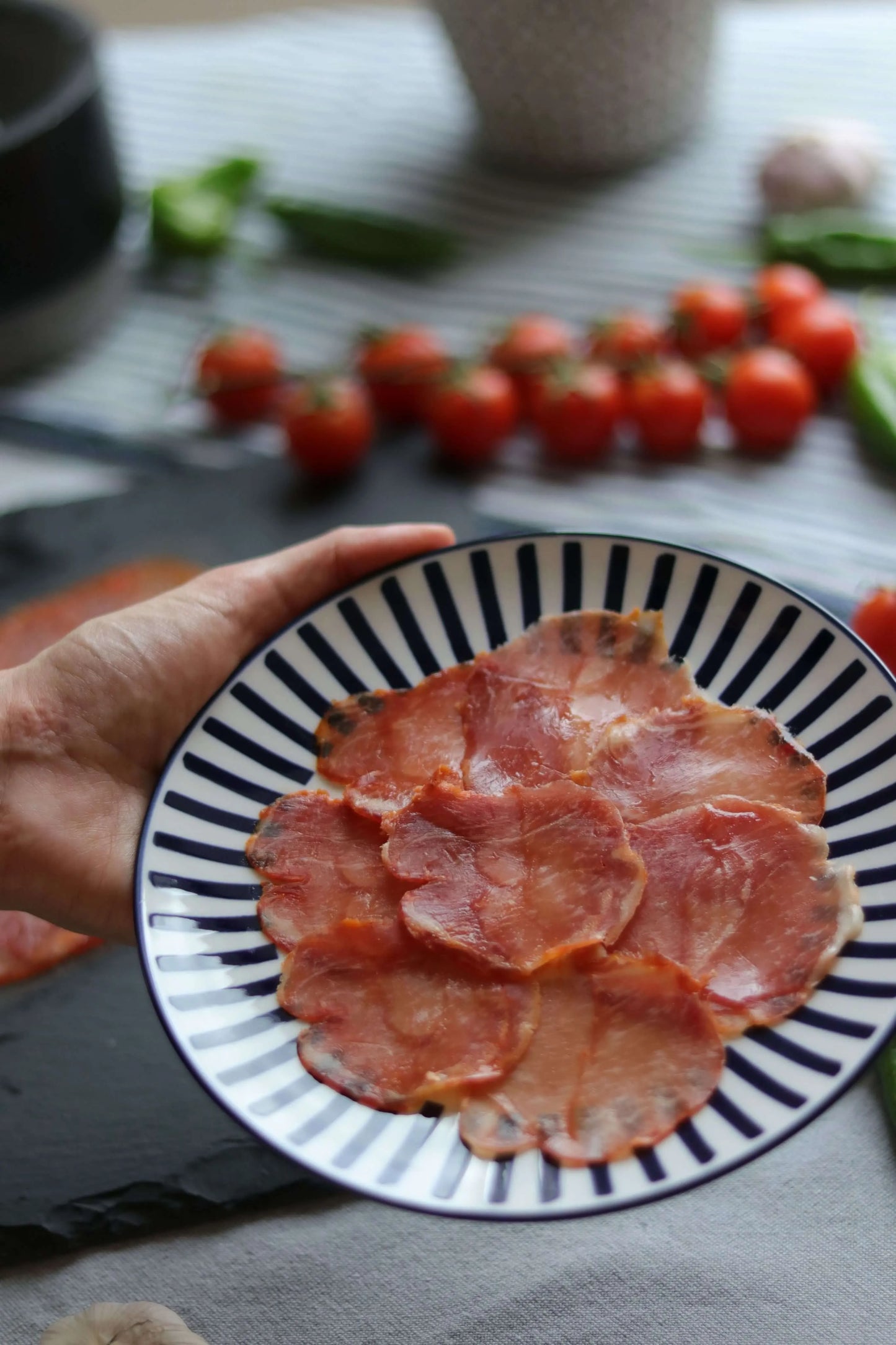 Teller mit dünn geschnittenen Schweinelende Ibérico (Lomo), serviert auf blau-weißem Teller, im Hintergrund frische Tomaten und Pimientos. 