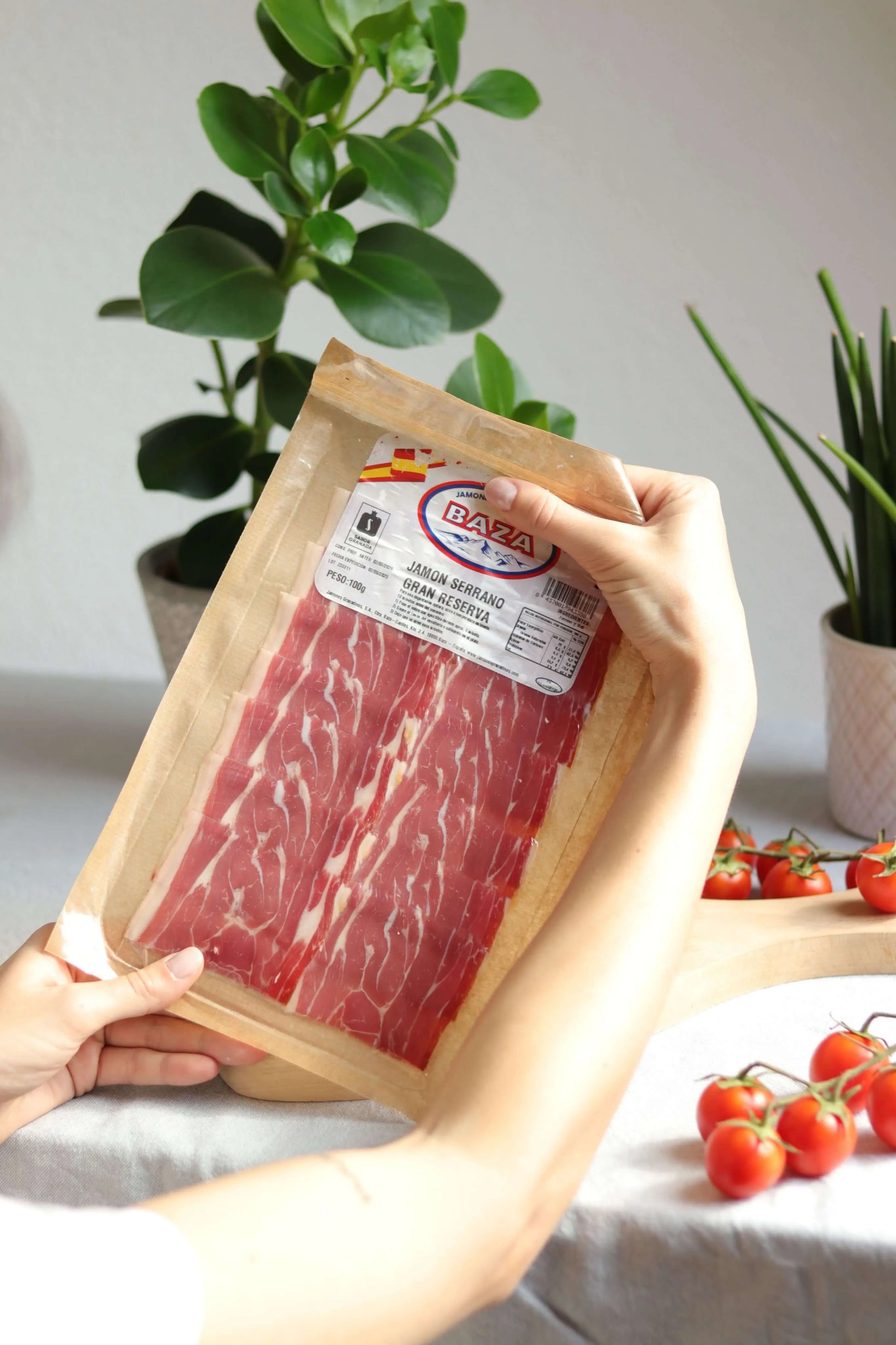 Vakuumverpackter Serrano Gran Reserva Schinken von Baza, dünn geschnitten, in den Händen gehalten mit Tomaten im Hintergrund.